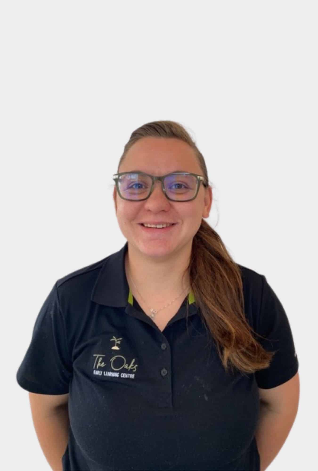 Person wearing glasses and a black polo shirt with "The Oaks Early Learning Centre" logo, smiling at the camera against a plain white background.