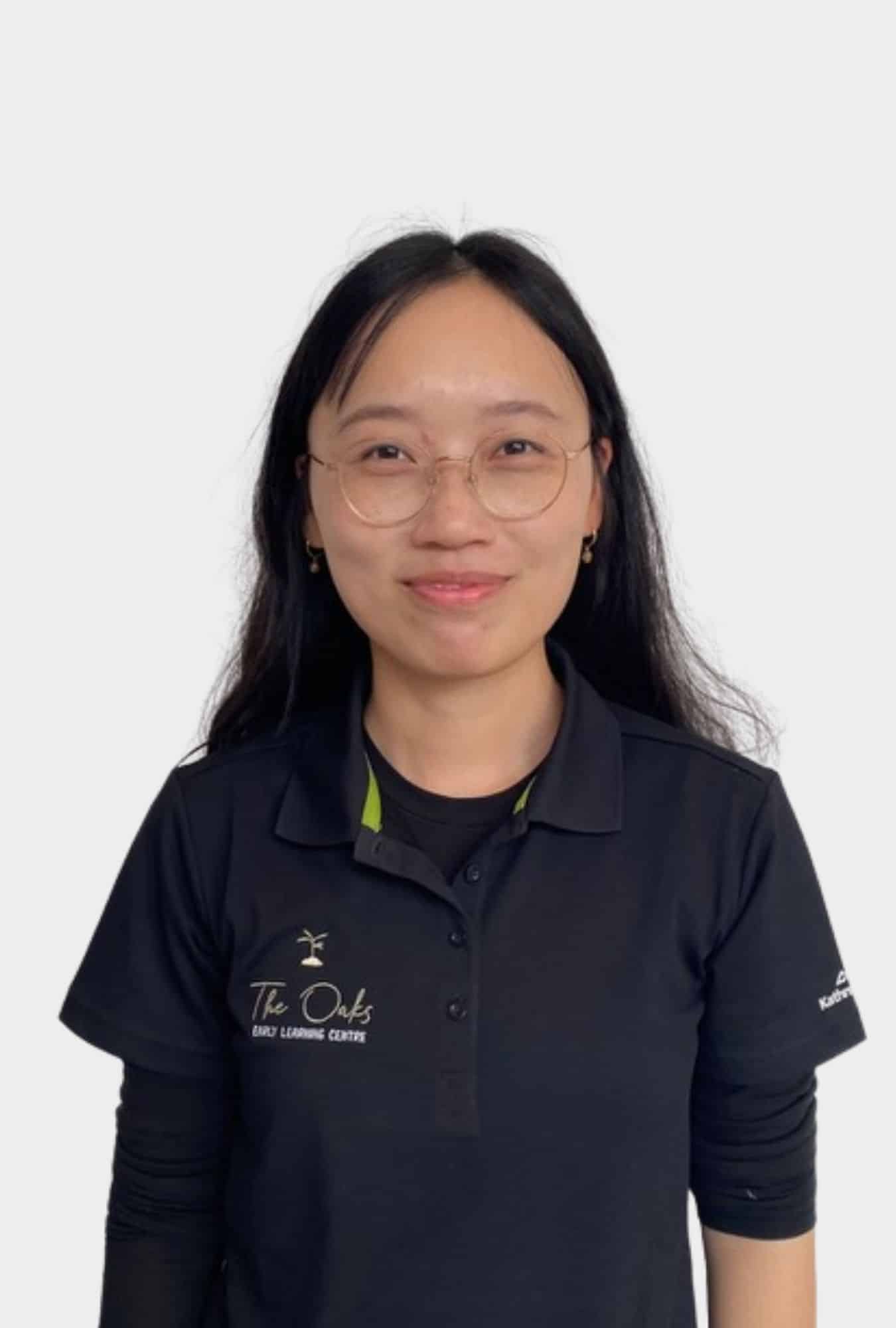 A woman with long black hair and glasses, wearing a black polo shirt with "The Oaks Early Learning Centre" logo, stands facing the camera against a plain background.